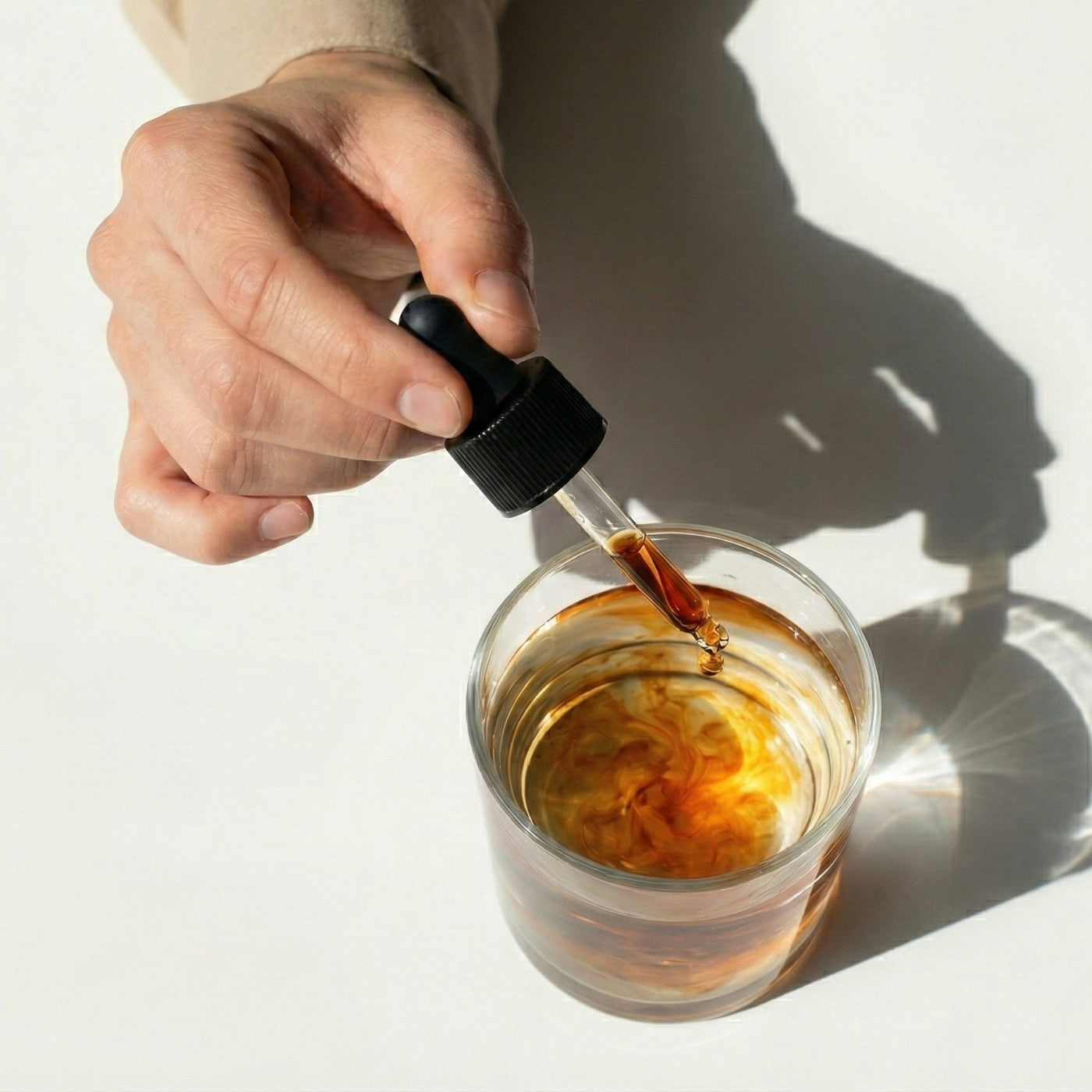 Hand using a dropper to polyextract mushroom liquid from a glass bottle into a glass of amber liquid on a light background